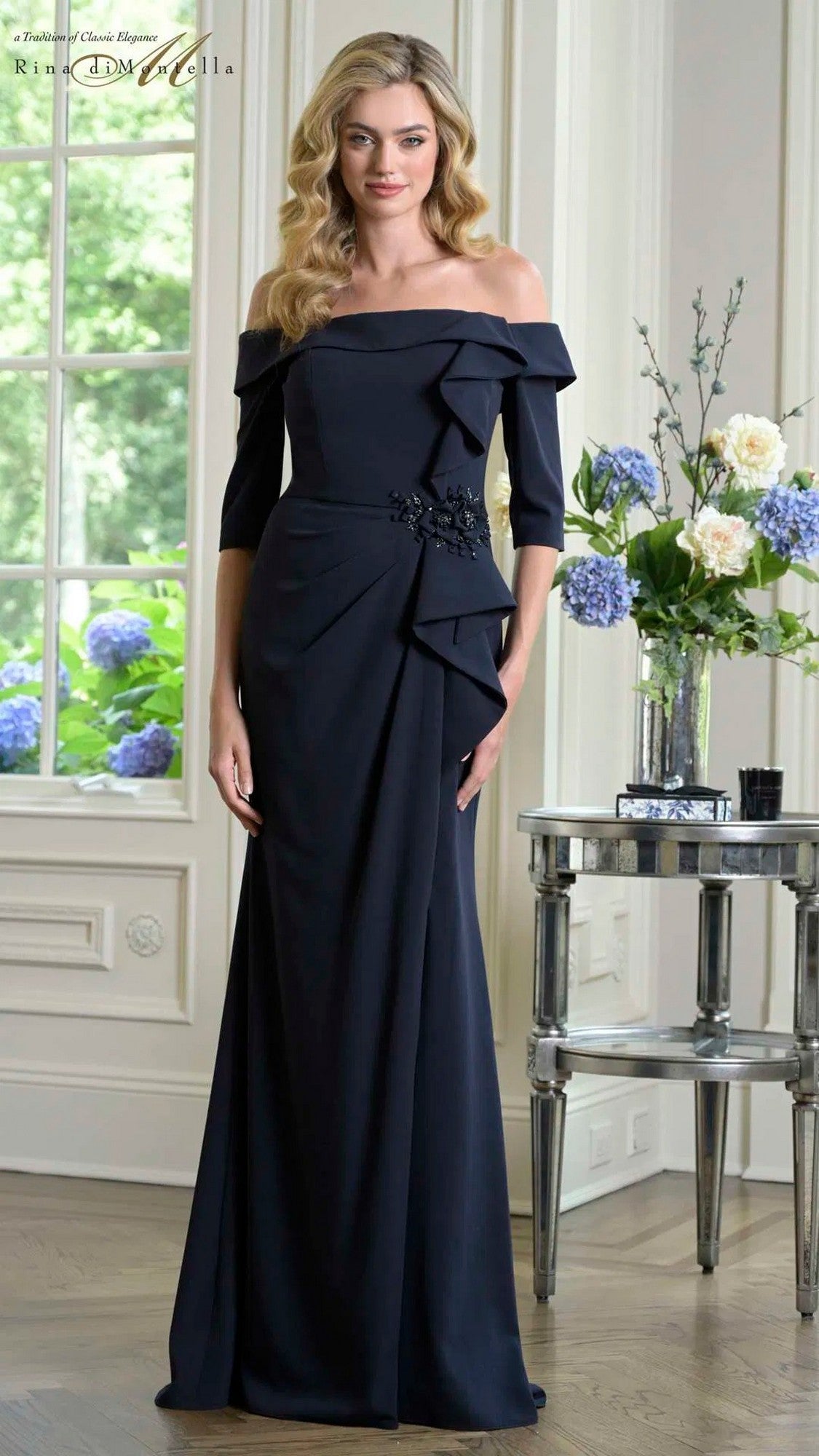 A woman in a navy Rina Di Montella RD3202 long formal dress stands indoors near a flower-adorned table, framed by tall windows with white trim.